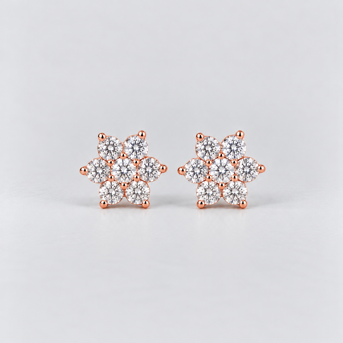 A pair of rose gold stud earrings featuring a snowflake-like cluster design set with clear round-cut gemstones, photographed on a light gray background.