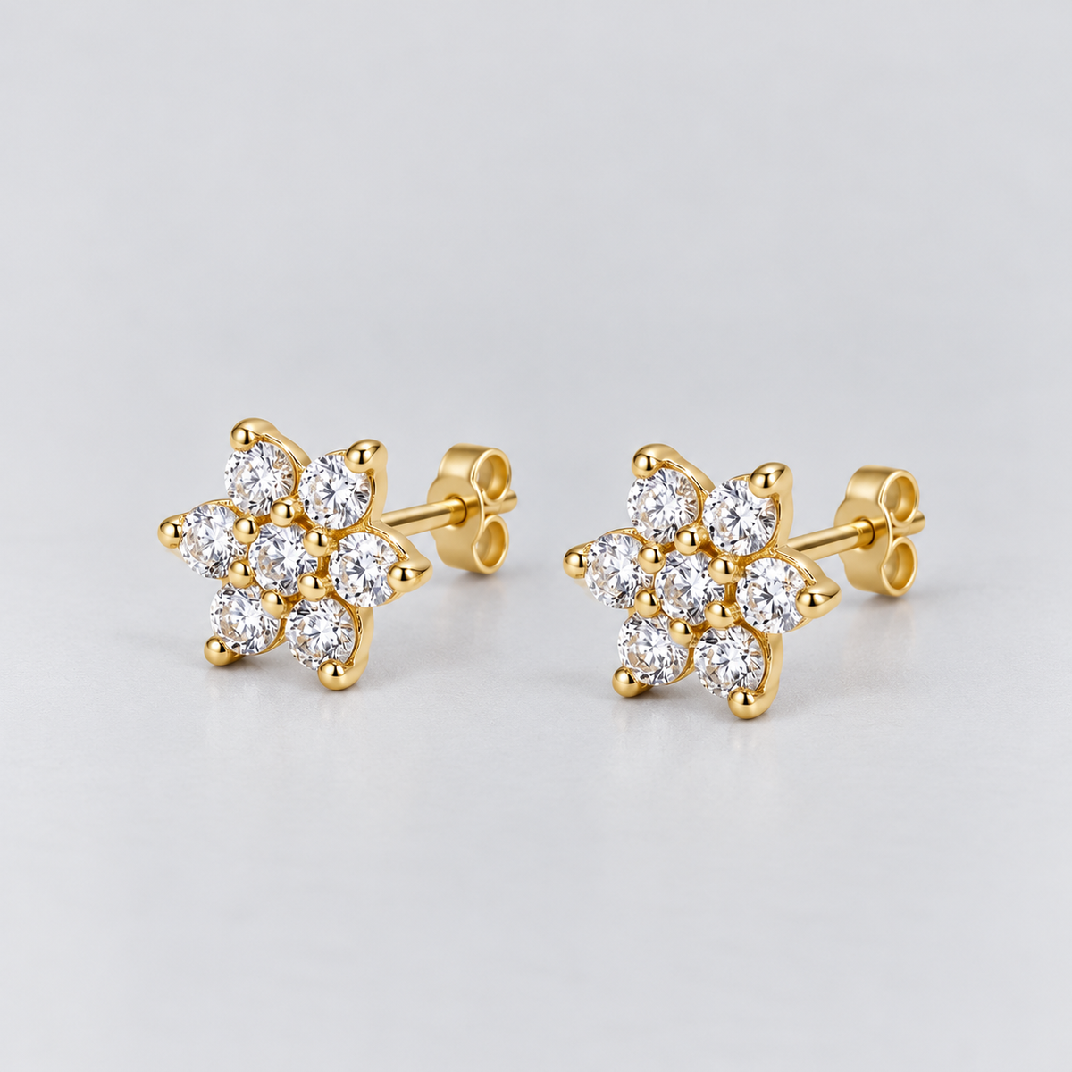 A pair of gold stud earrings featuring a cluster of round-cut clear gemstones in a flower-like arrangement, set on a light gray background.