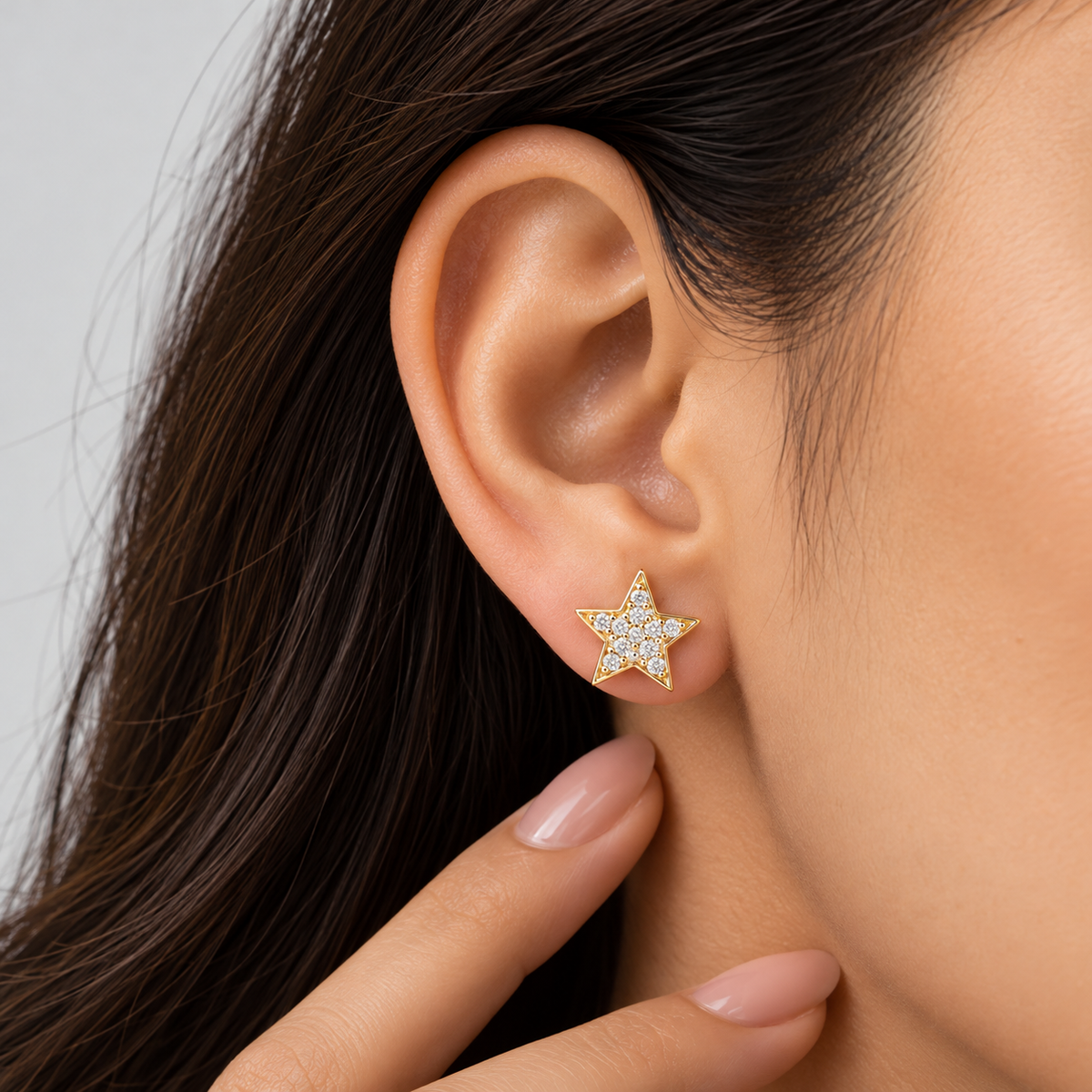 A close-up of a model's ear wearing a gold star-shaped stud earring set with small clear crystals, with her hand resting near her neck.