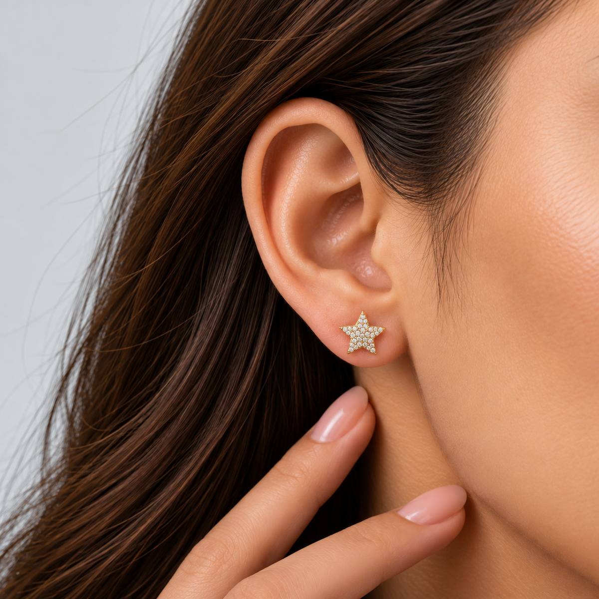 A close-up of a woman's ear wearing a small gold star stud earring set with clear crystals, with her hand resting near her neck against a light gray background.