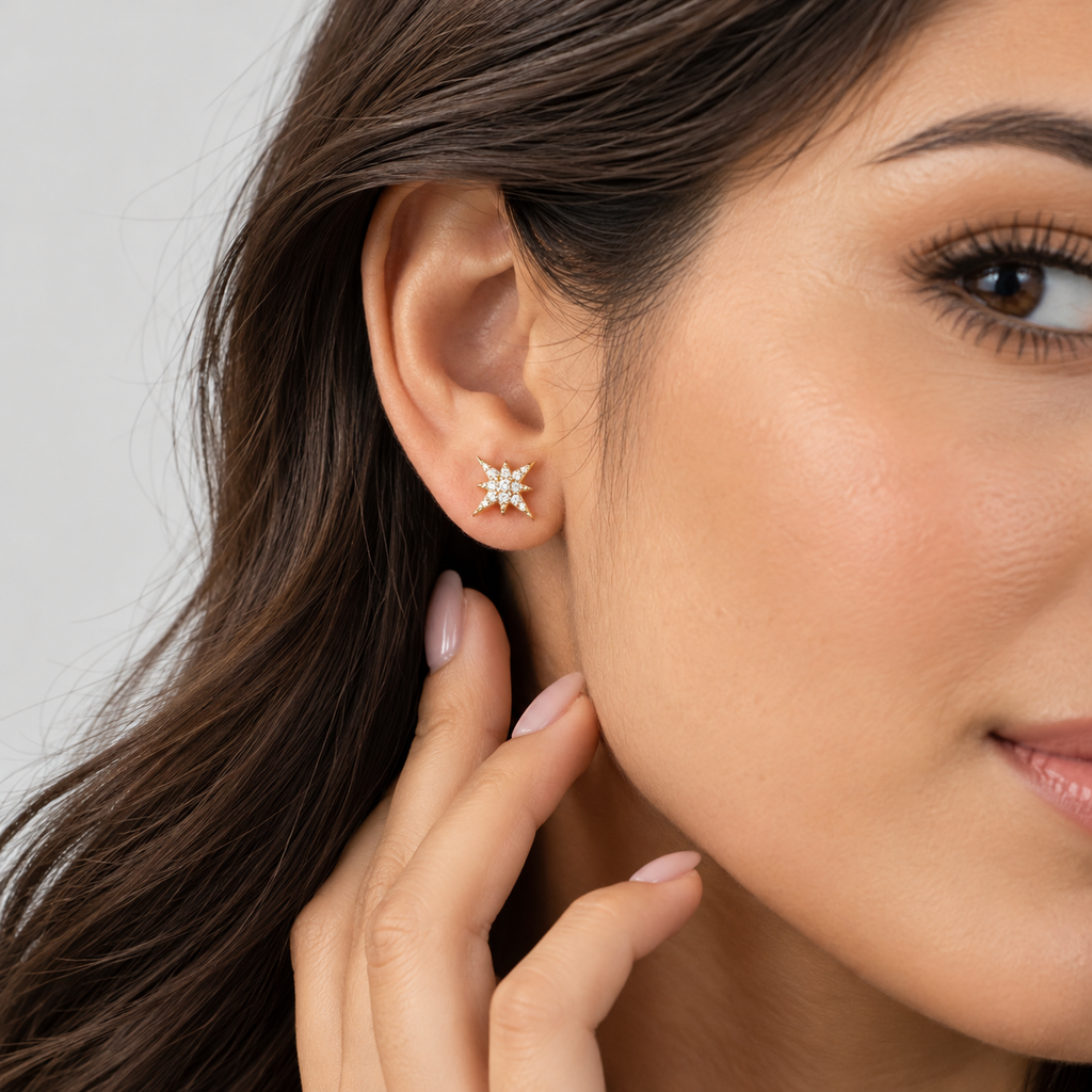 A close-up of a woman's ear wearing a small star-shaped stud earring with clear stones in a silver-toned setting, against a soft light gray background.
