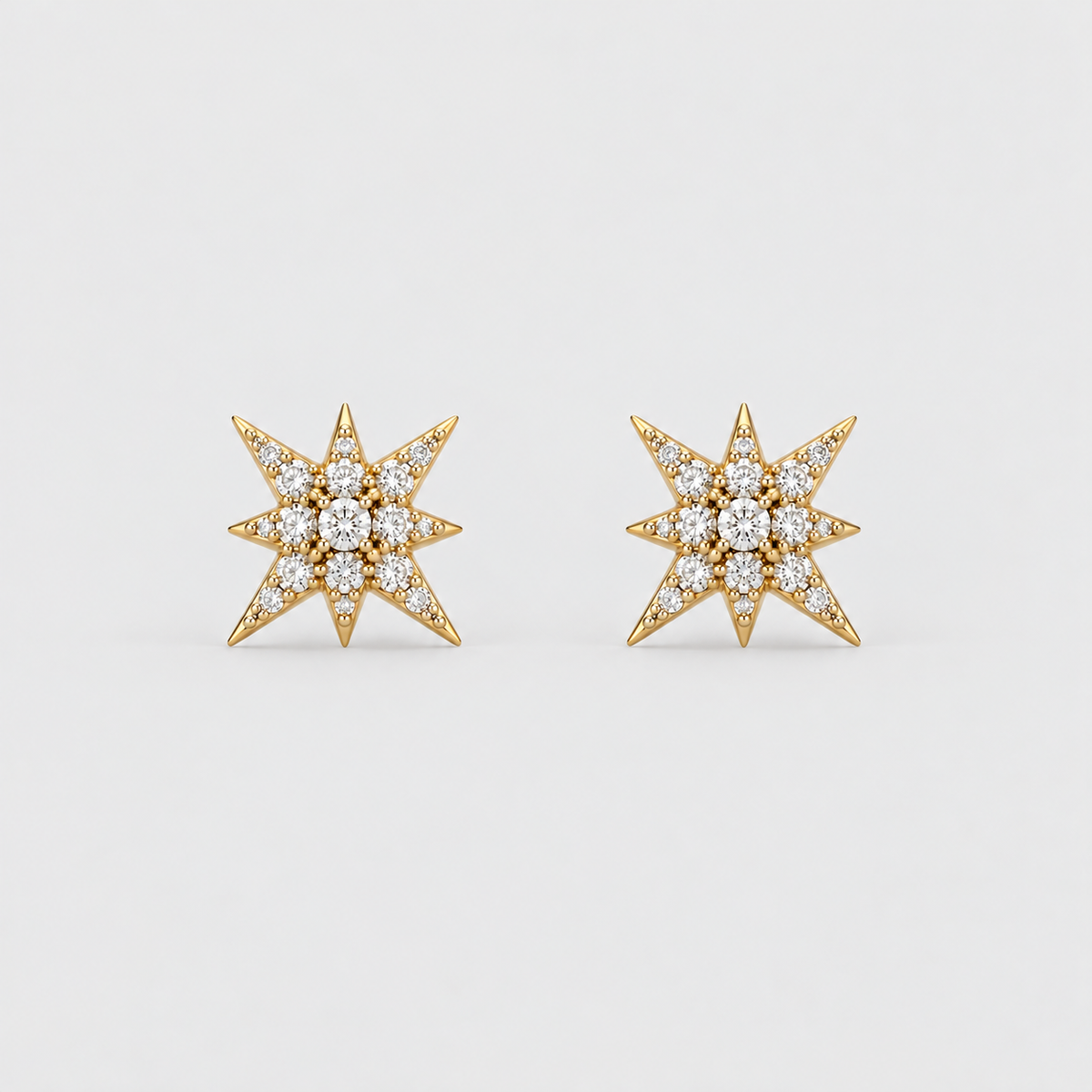 A pair of gold starburst stud earrings set with clear pavé gemstones, shown on a light gray background.