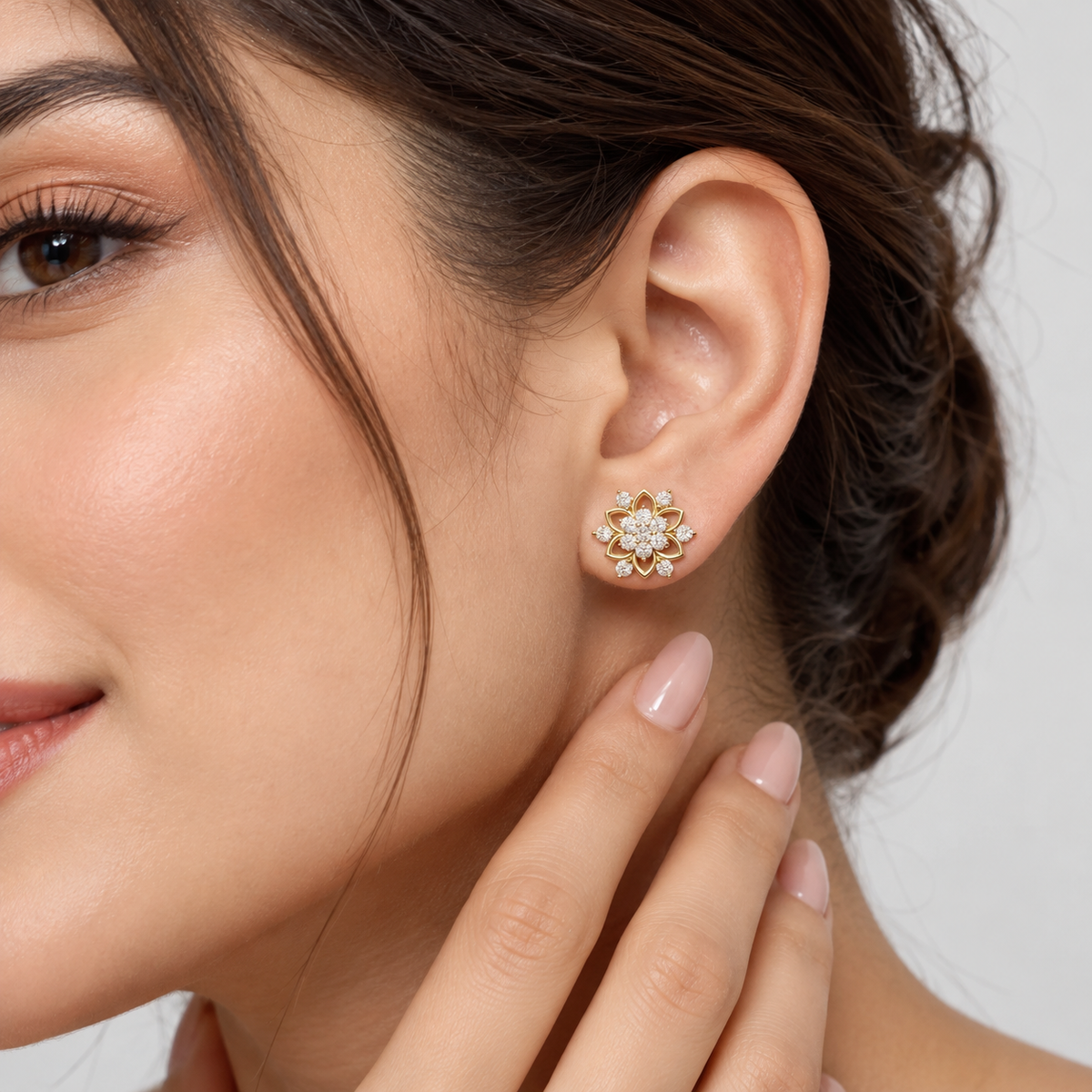 A close-up of a woman's ear wearing a small gold flower-shaped stud earring with clear gemstones, against a soft light gray background.