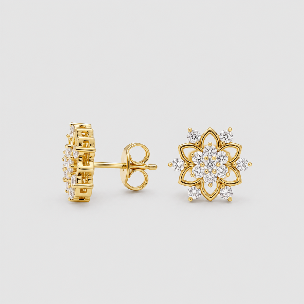 A pair of gold stud earrings on a light gray background. One earring is shown from the side/back view, displaying the post and butterfly back, while the other is shown from the front, featuring a flower-like design with clear gemstones.