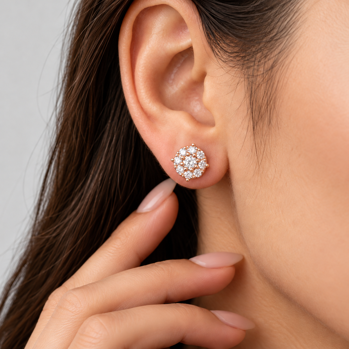 A close-up of a woman's ear wearing a small cluster stud earring with clear stones in a gold-tone setting, with her hand near her neck against a light gray background.