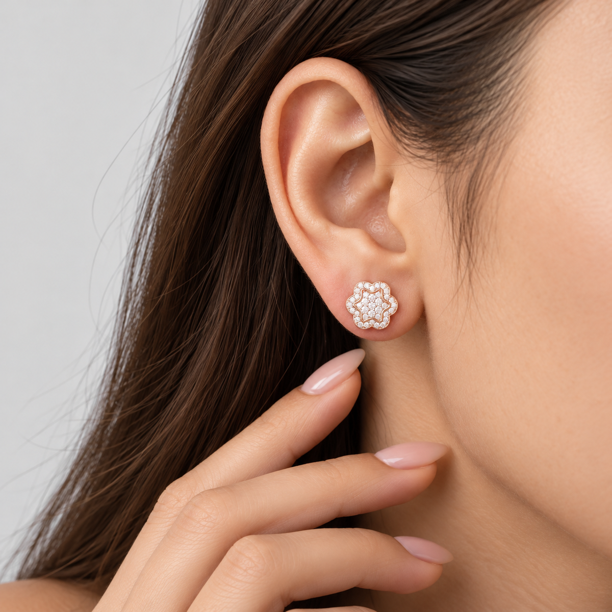 A close-up of a woman's ear wearing a small flower-shaped stud earring with clear stones in a silver-toned setting, against a soft light gray background.