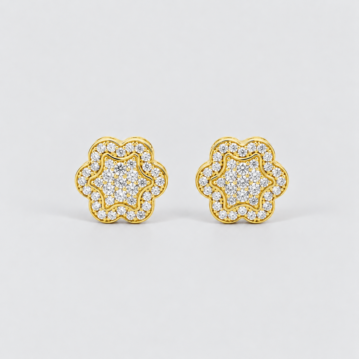 A pair of gold flower-shaped stud earrings paved with clear round stones, set against a clean light gray background.