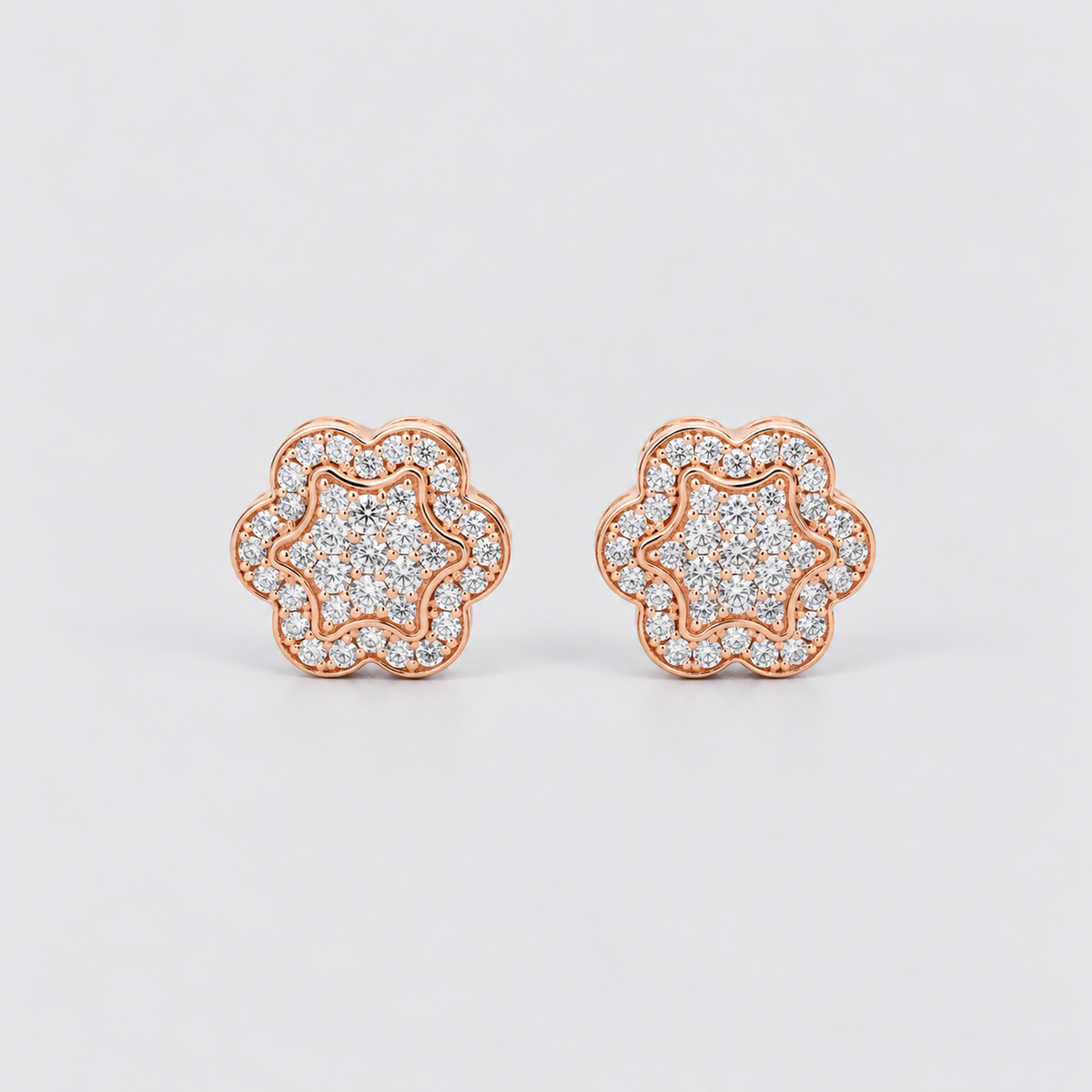 A pair of rose gold stud earrings with a double halo design, featuring pavé-set clear stones. The earrings are shown on a clean, light gray background.
