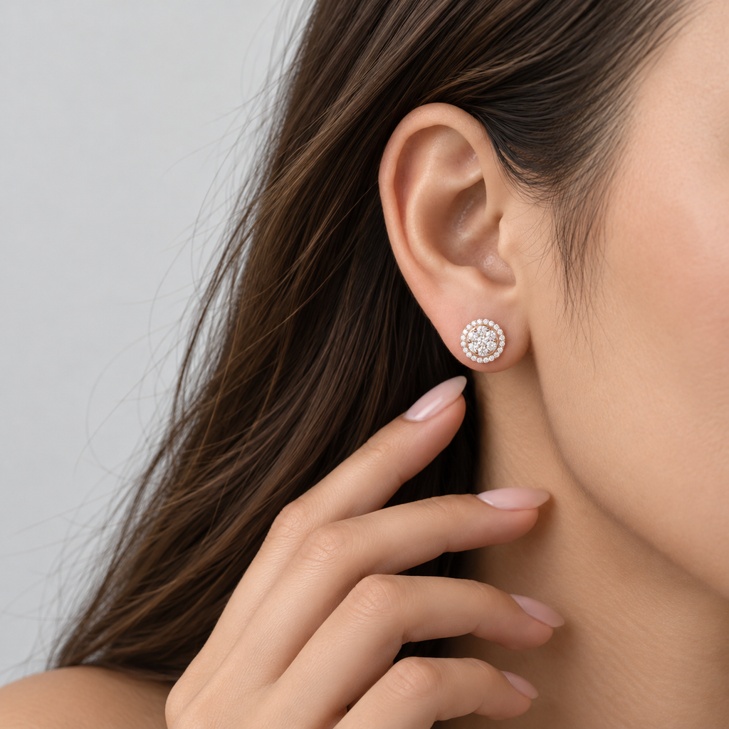 A close-up of a woman's ear wearing a small round stud earring with a halo of clear stones in a silver-toned setting, with her hand near her neck against a soft light gray background.