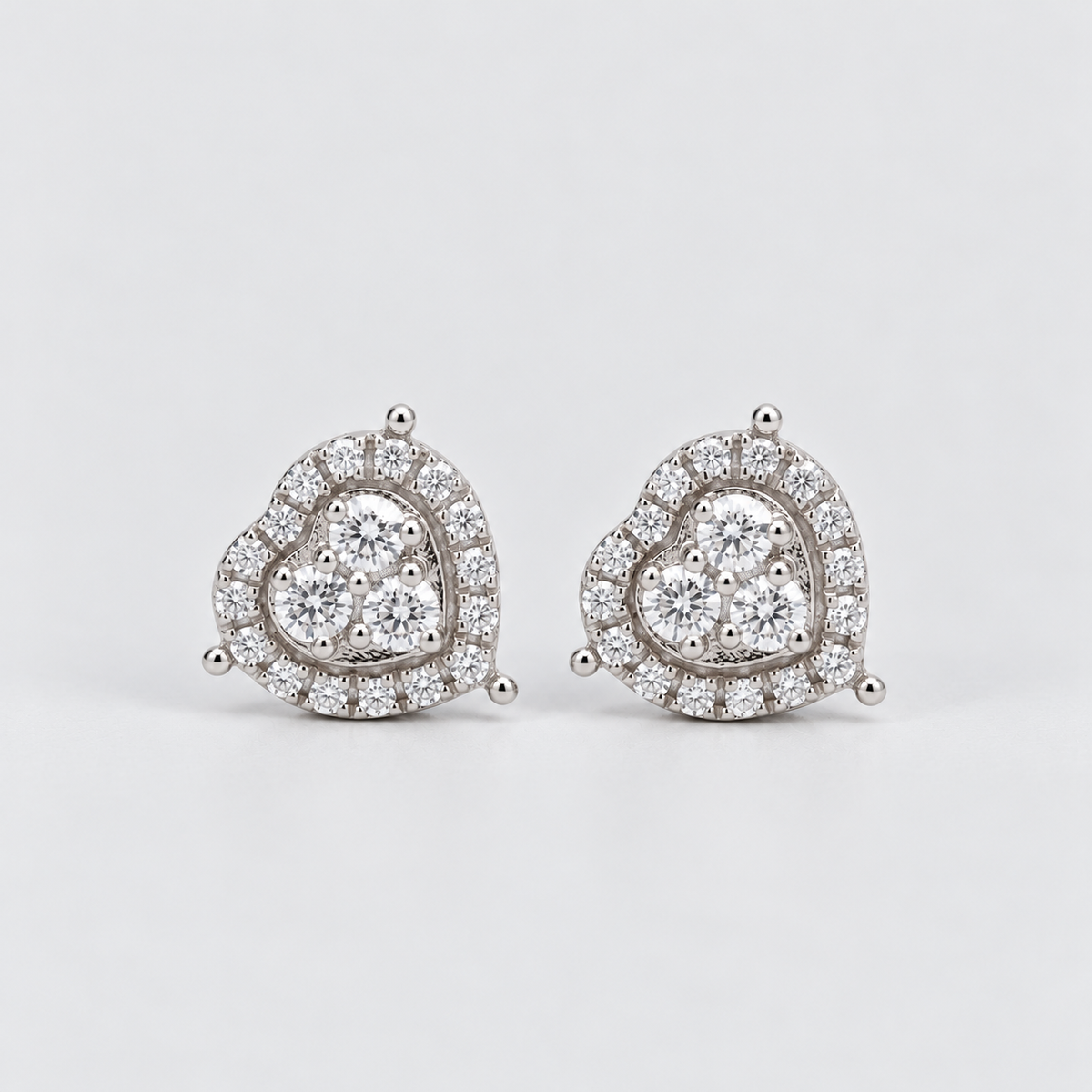 A pair of heart-shaped stud earrings in silver-tone metal, adorned with clear pavé diamonds and a central cluster of round diamonds, set against a plain light gray background.