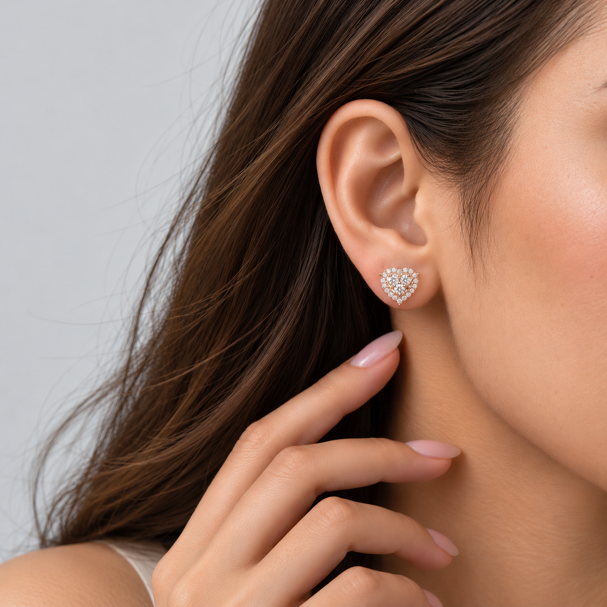 A close-up of a woman's ear wearing a small heart-shaped stud earring made of clear stones in a silver-toned setting, with her hand near her neck against a light gray background.