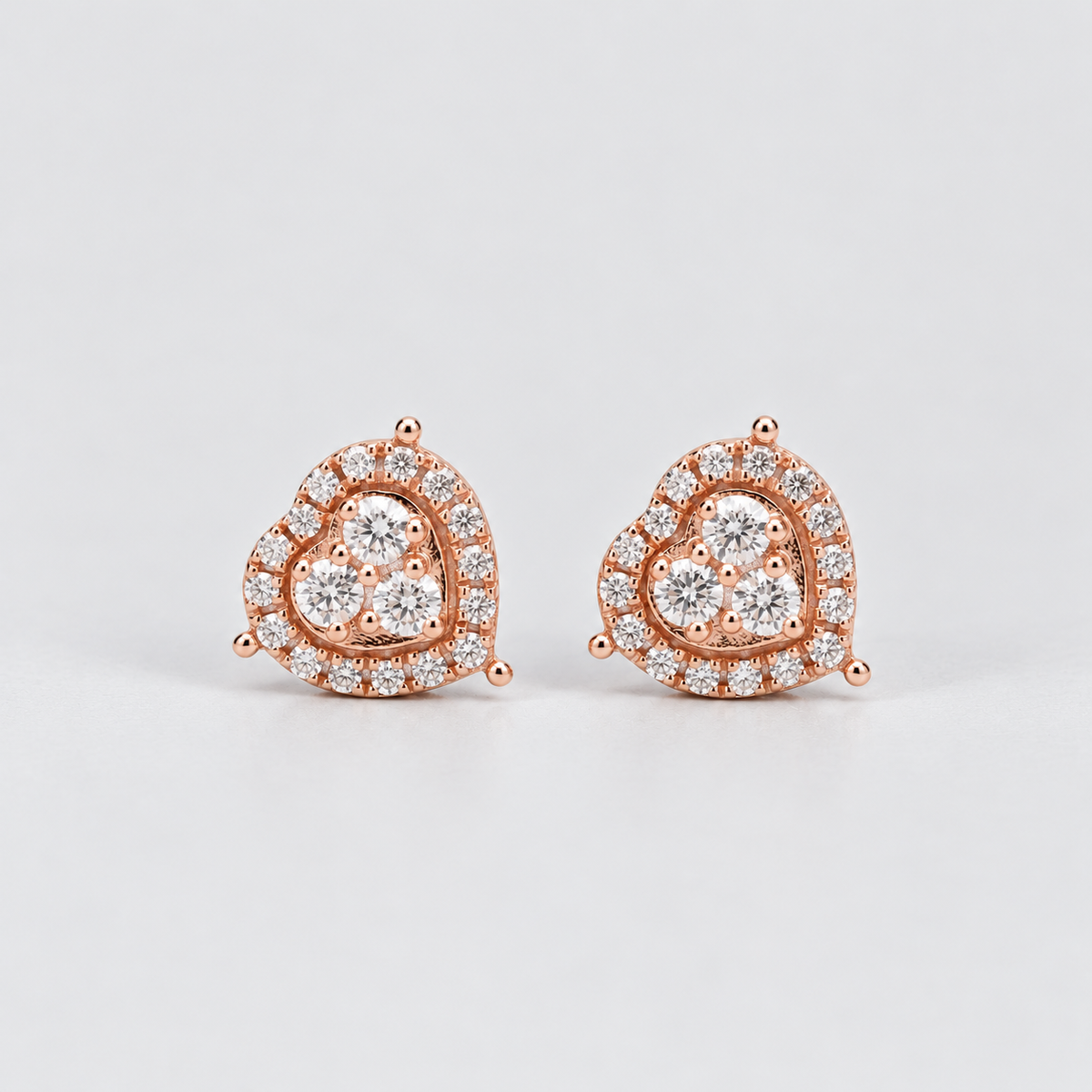 A pair of rose gold heart-shaped stud earrings set with clear round-cut gemstones, photographed on a clean light gray background.