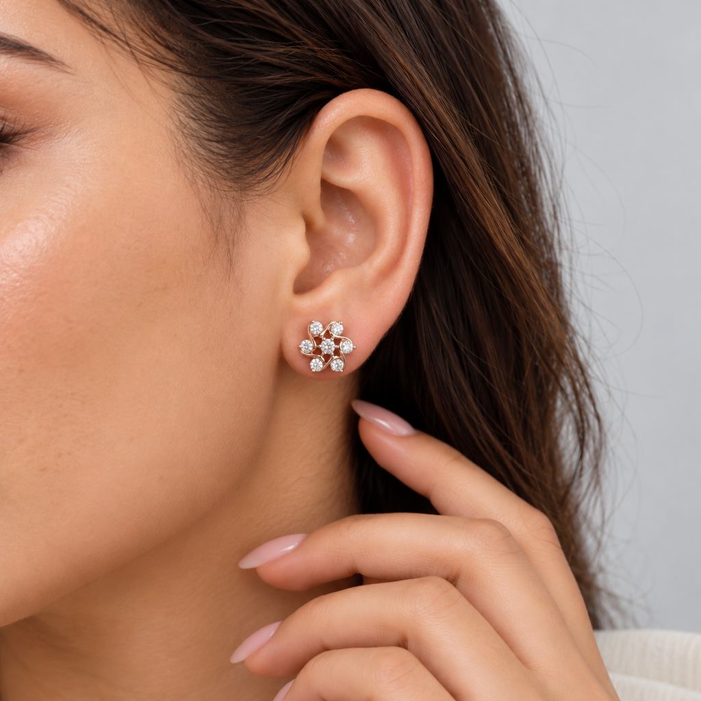 A close-up of a woman's ear wearing a small flower-shaped stud earring with clear gemstones in a silver-toned setting, against a soft light gray background.