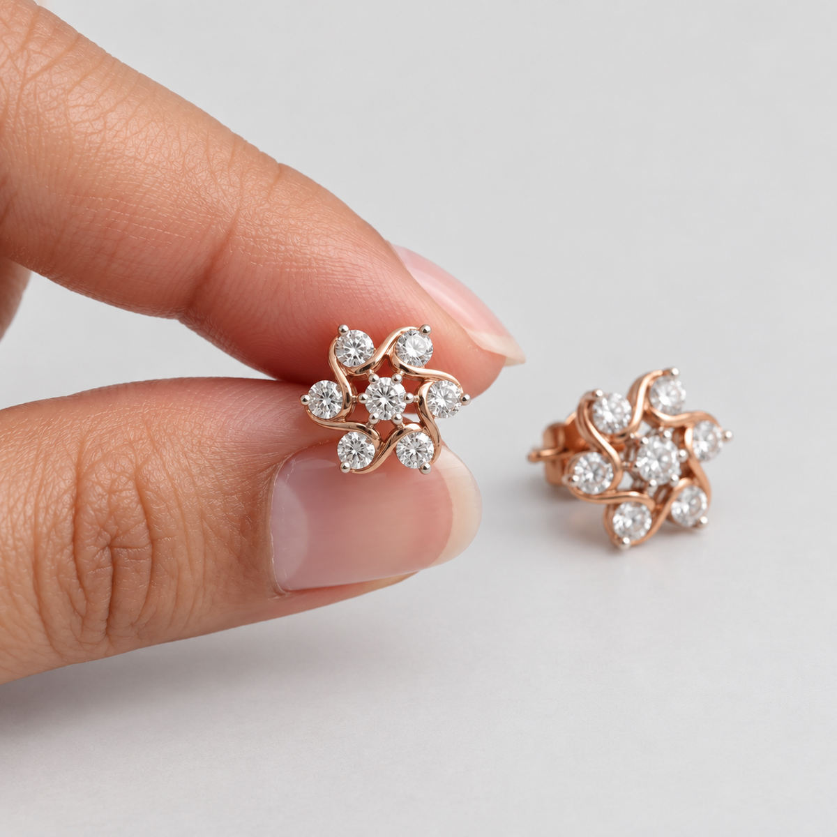 Rose gold flower-shaped stud earrings with clear gemstones, shown with one earring held between fingers and the other resting on a light gray background.