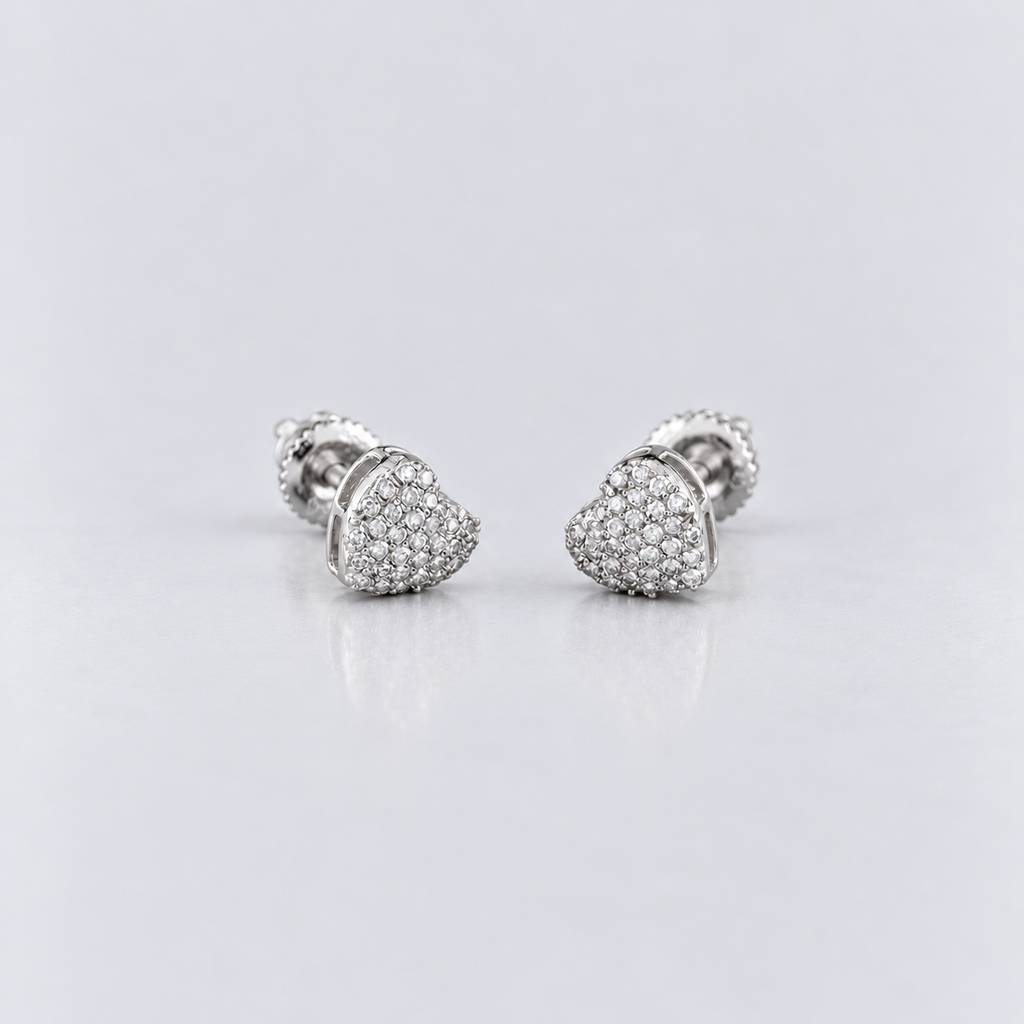 A pair of silver-toned heart-shaped stud earrings paved with small clear stones, displayed on a clean light gray reflective surface.