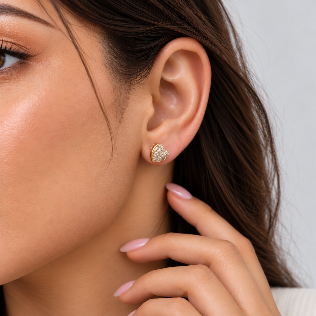 A close-up of a woman's ear wearing a small gold heart-shaped stud earring adorned with tiny clear pavé stones, against a soft light gray background.