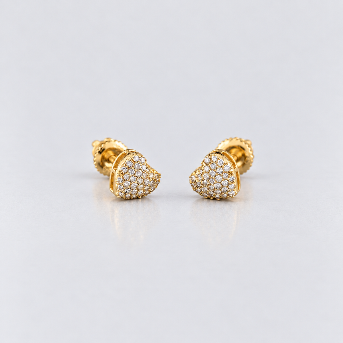 A pair of gold heart-shaped stud earrings paved with small clear stones, displayed on a light gray reflective surface.