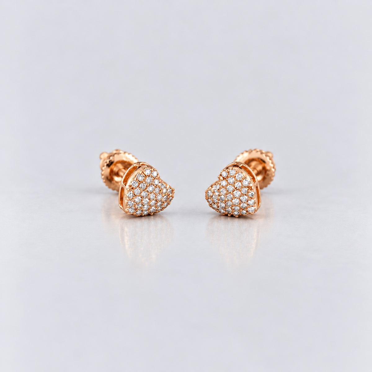 A pair of rose gold heart-shaped stud earrings paved with small clear stones, displayed on a light gray reflective surface.