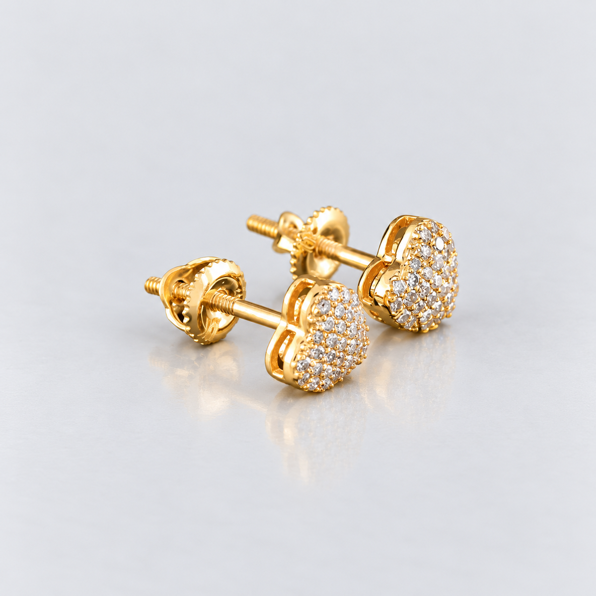 A pair of gold heart-shaped stud earrings adorned with pavé-set clear stones, shown on a clean light gray reflective surface.