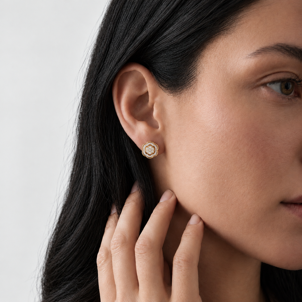 A close-up of a woman wearing a small gold stud earring with a halo of clear stones, shown against a clean light gray background.
