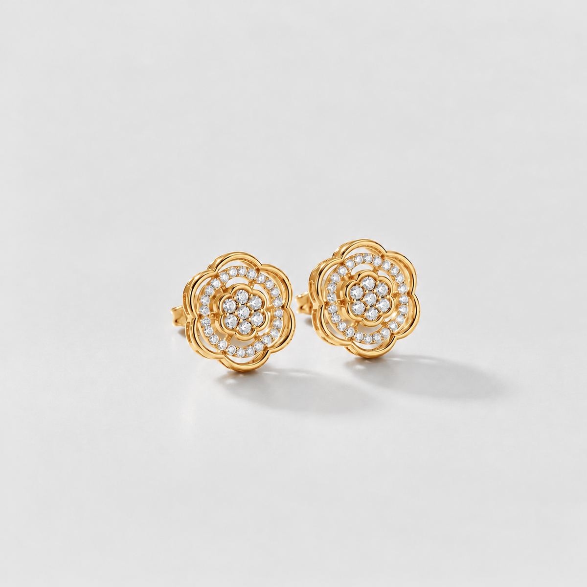 A pair of gold flower-shaped stud earrings adorned with small clear pavé stones, photographed on a clean light gray background.