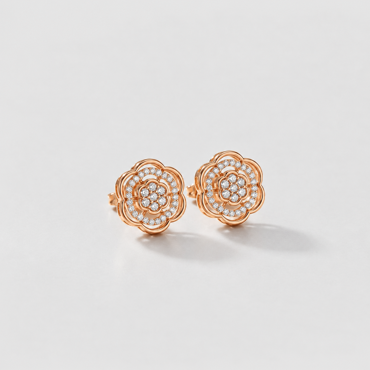 A pair of rose gold stud earrings with a layered flower design, featuring small clear pavé gemstones in the center, set against a clean light gray background.