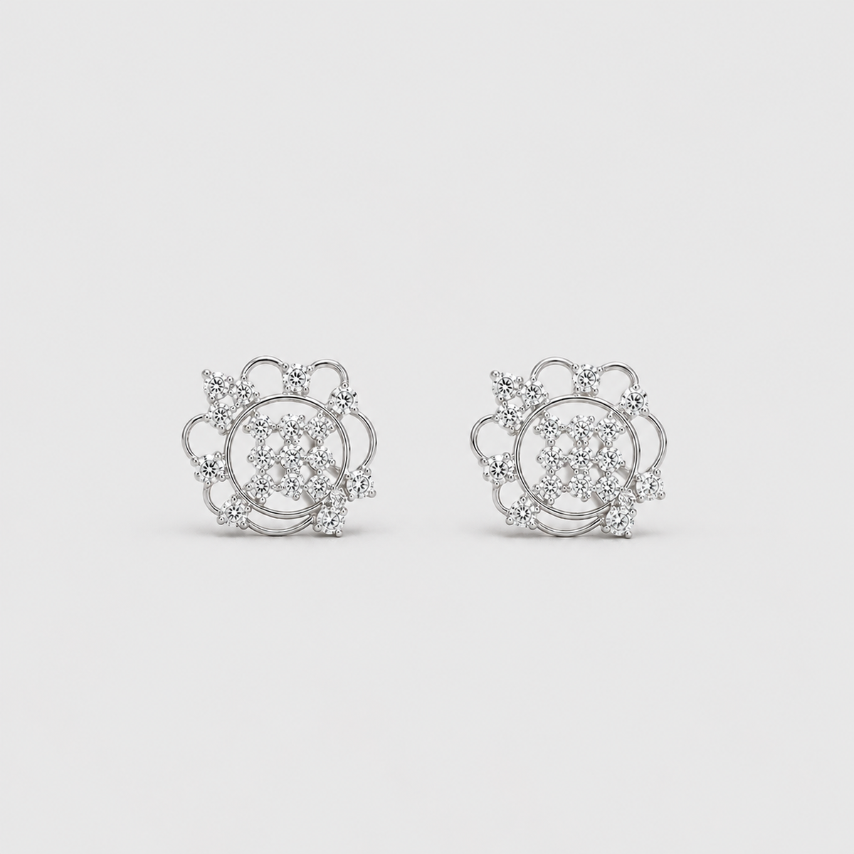 A pair of silver-toned stud earrings with intricate circular and floral designs set with clear gemstones, displayed on a light gray background.
