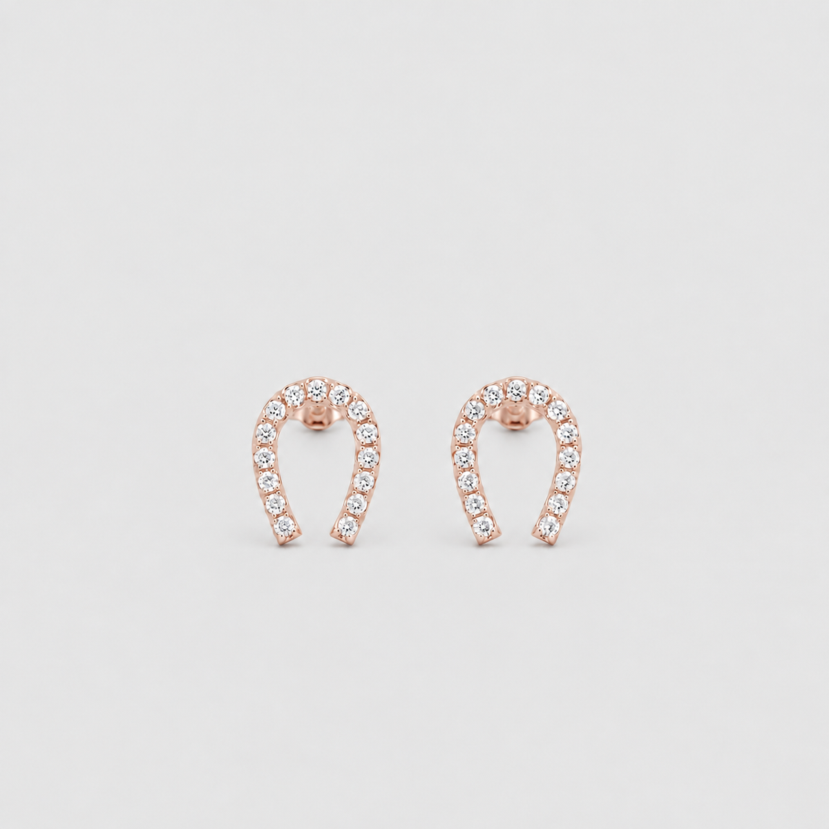 A pair of rose gold horseshoe-shaped stud earrings set with small clear stones, displayed on a light gray background.