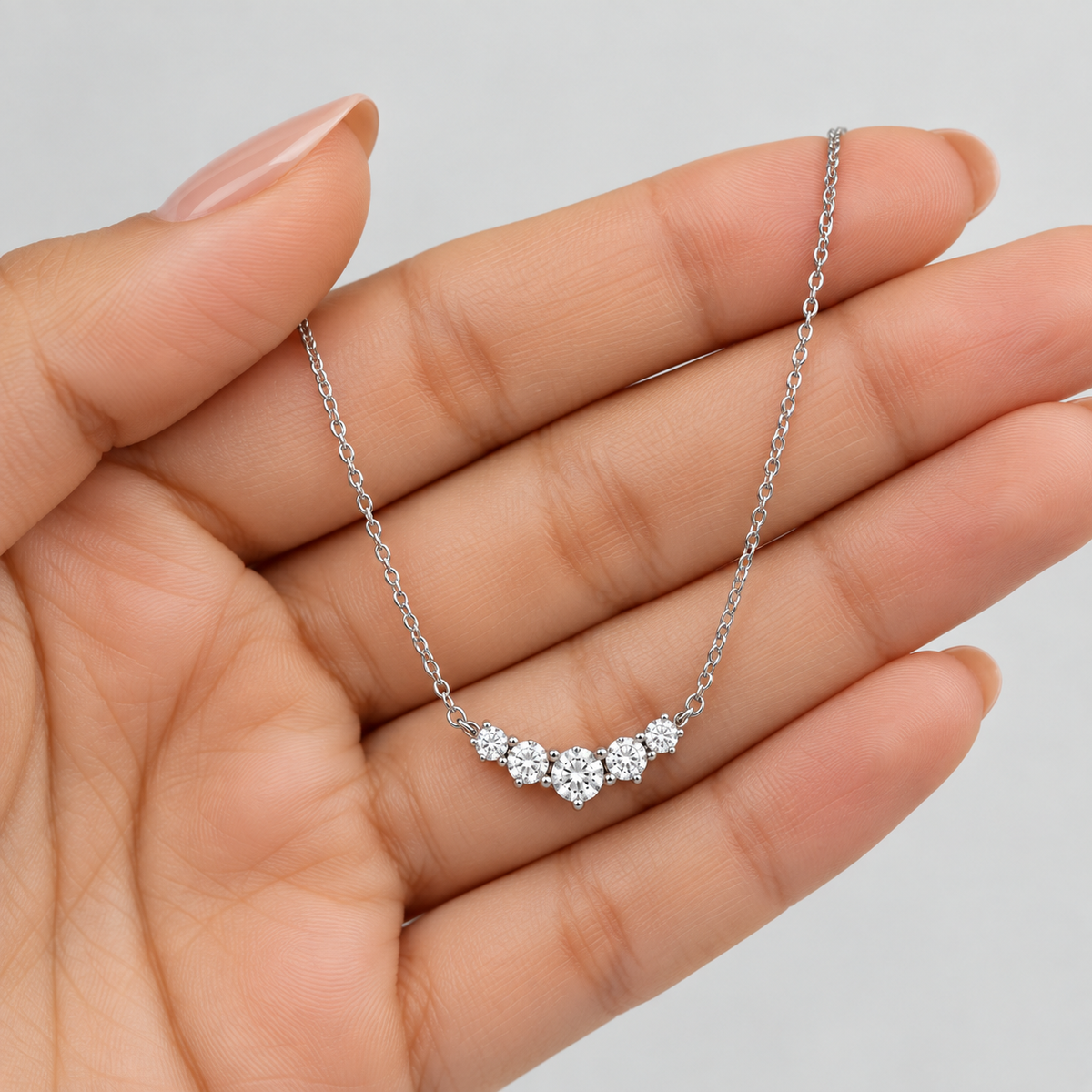 A delicate silver chain necklace with a small cluster of clear round-cut gemstones, held in the palm of a hand against a light gray background.