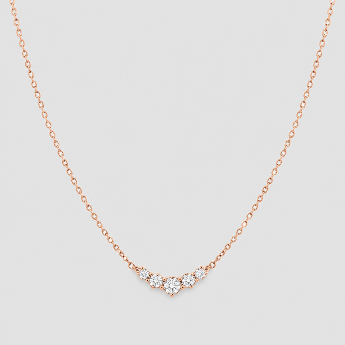 A delicate rose gold chain necklace with a small curved cluster of clear gemstones at the center, displayed against a light gray background.