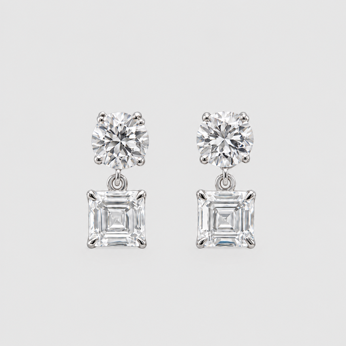 A pair of silver-tone drop earrings featuring a round brilliant-cut clear gemstone on top and a square baguette-cut clear gemstone below, set against a light gray background.