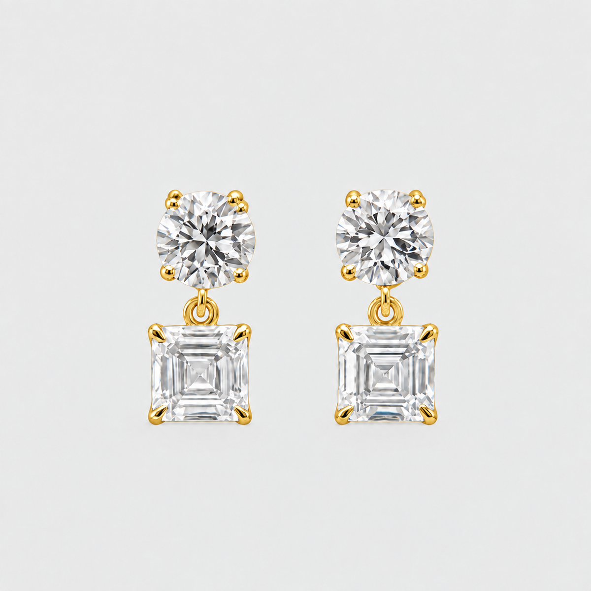 A pair of gold drop earrings featuring a round brilliant-cut clear gemstone on top and a larger square baguette-cut clear gemstone below, set in yellow gold prongs against a light gray background.