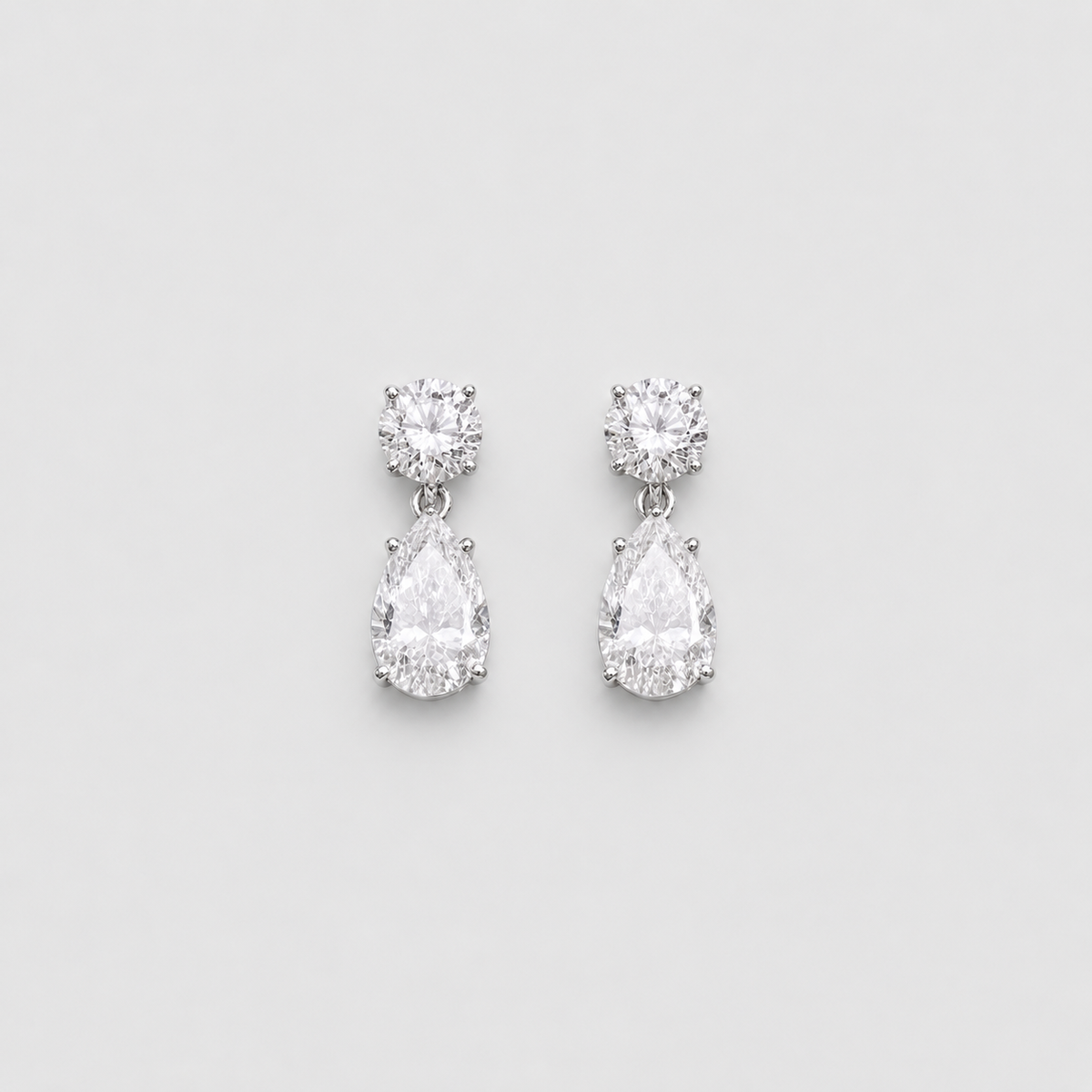 A pair of silver drop earrings featuring a round-cut clear gemstone at the top and a pear-cut clear gemstone below, set against a light gray background.