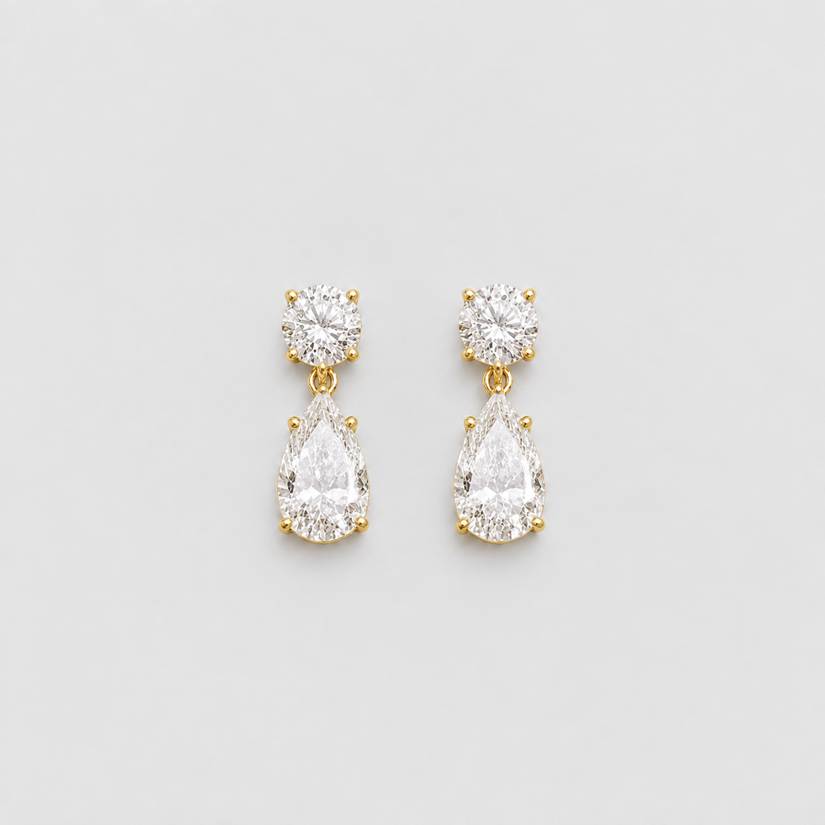 A pair of gold drop earrings featuring a round-cut clear gemstone in a prong setting at the top and a pear-cut clear gemstone below, displayed on a light gray background.