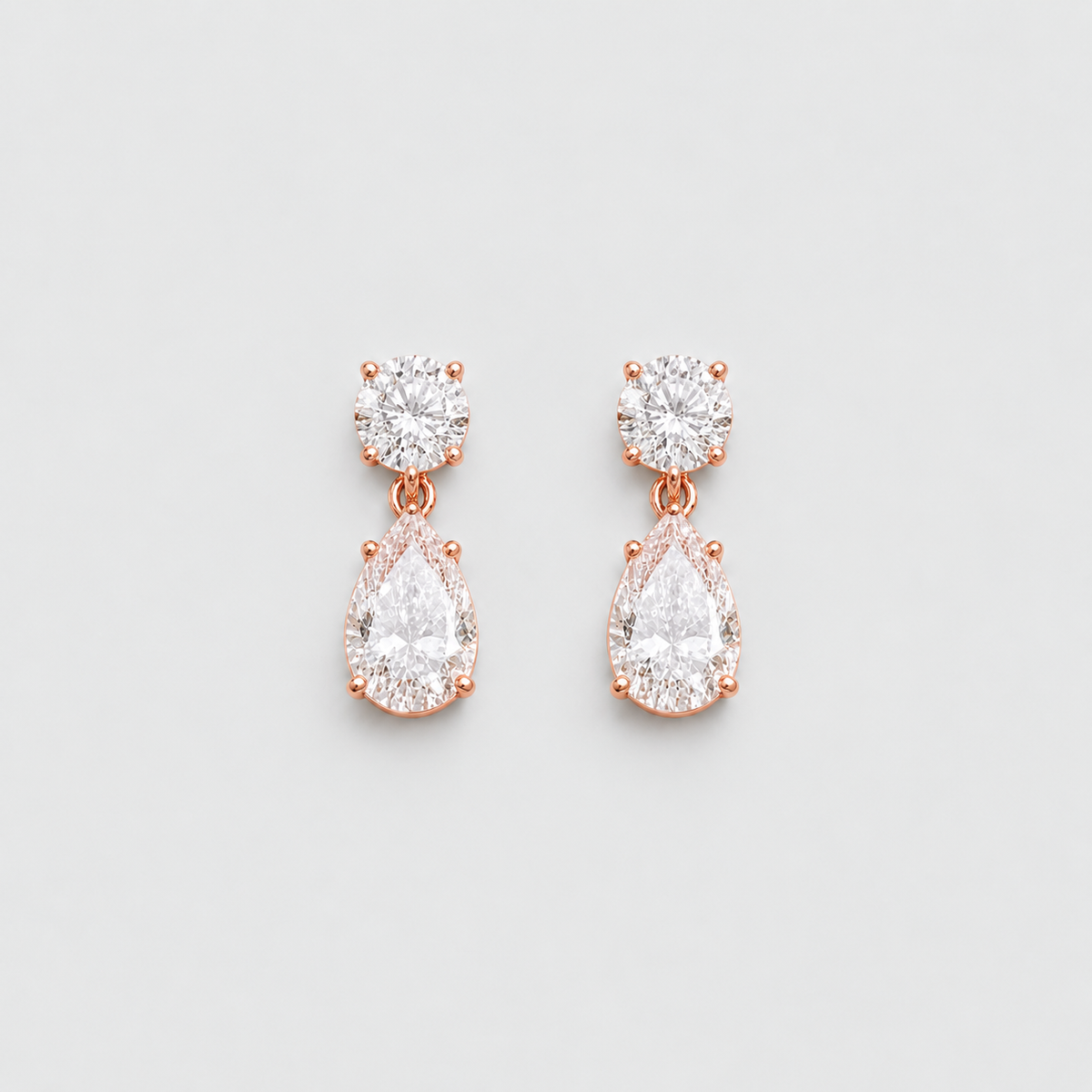 A pair of rose gold drop earrings with round and pear-cut clear gemstones, displayed on a clean light gray background.