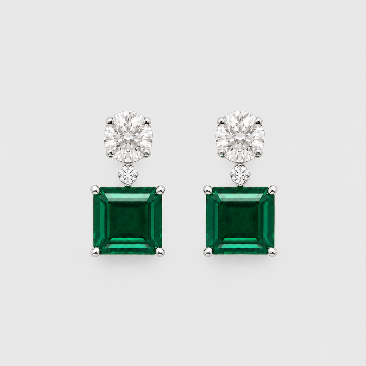 A pair of silver drop earrings featuring a square-cut emerald green gemstone and a round-cut clear gemstone in a silver setting, shown against a light gray background.