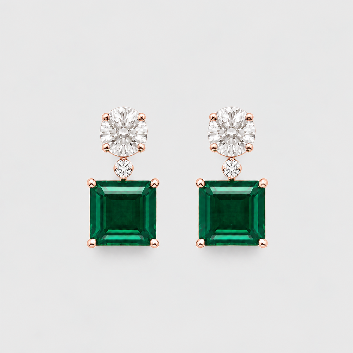 A pair of rose gold drop earrings featuring a large emerald-cut green gemstone and a round-cut clear gemstone above it, set against a light gray background.