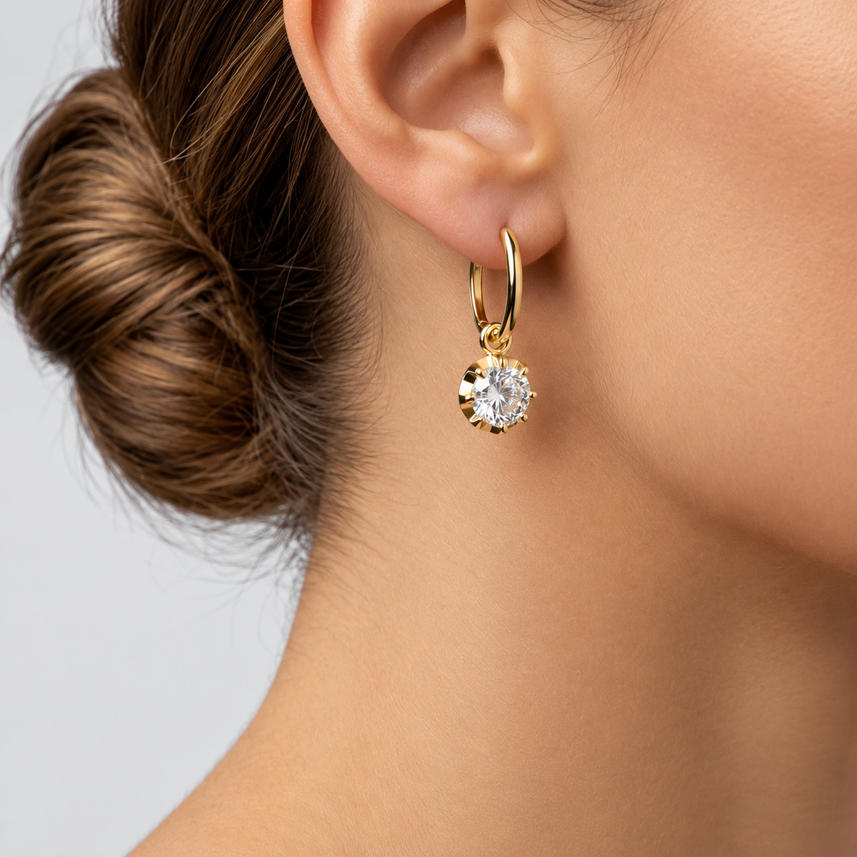 A close-up of a model's ear wearing a delicate gold huggie hoop earring with a heart-shaped clear gemstone pendant.