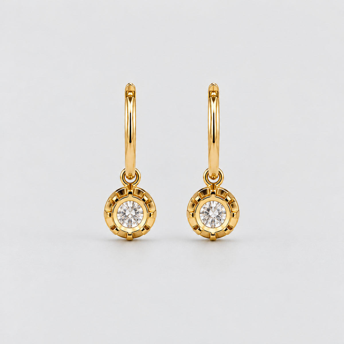 A pair of gold huggie hoop earrings with round bezel-set clear gemstones, displayed on a light gray background.