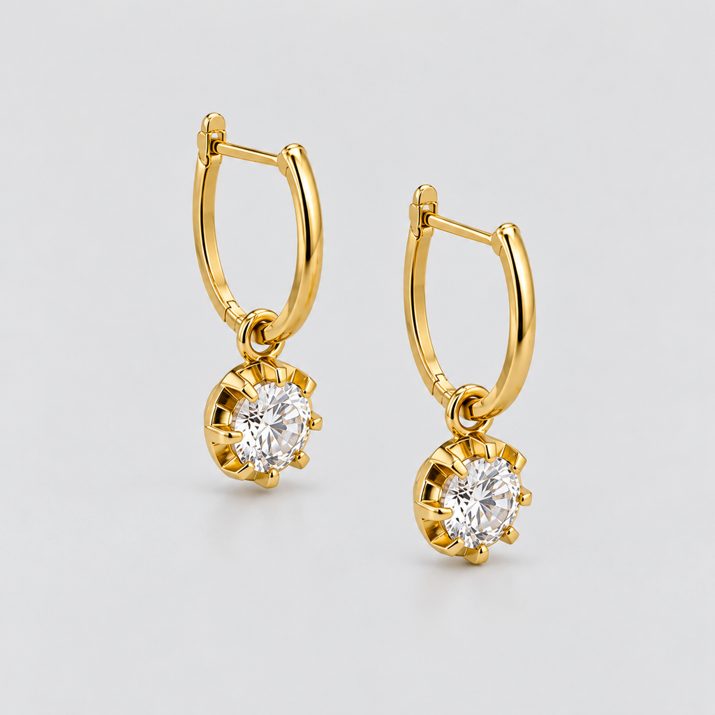 A pair of gold huggie hoop earrings with round-cut clear gemstones in a prong setting, displayed on a light gray background.