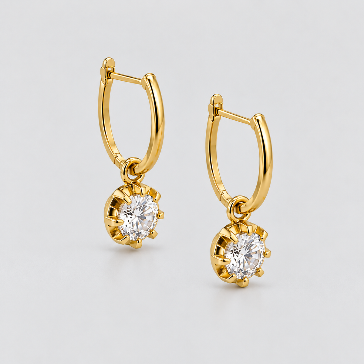A pair of gold huggie hoop earrings with round-cut clear gemstones in a prong setting, displayed on a light gray background.