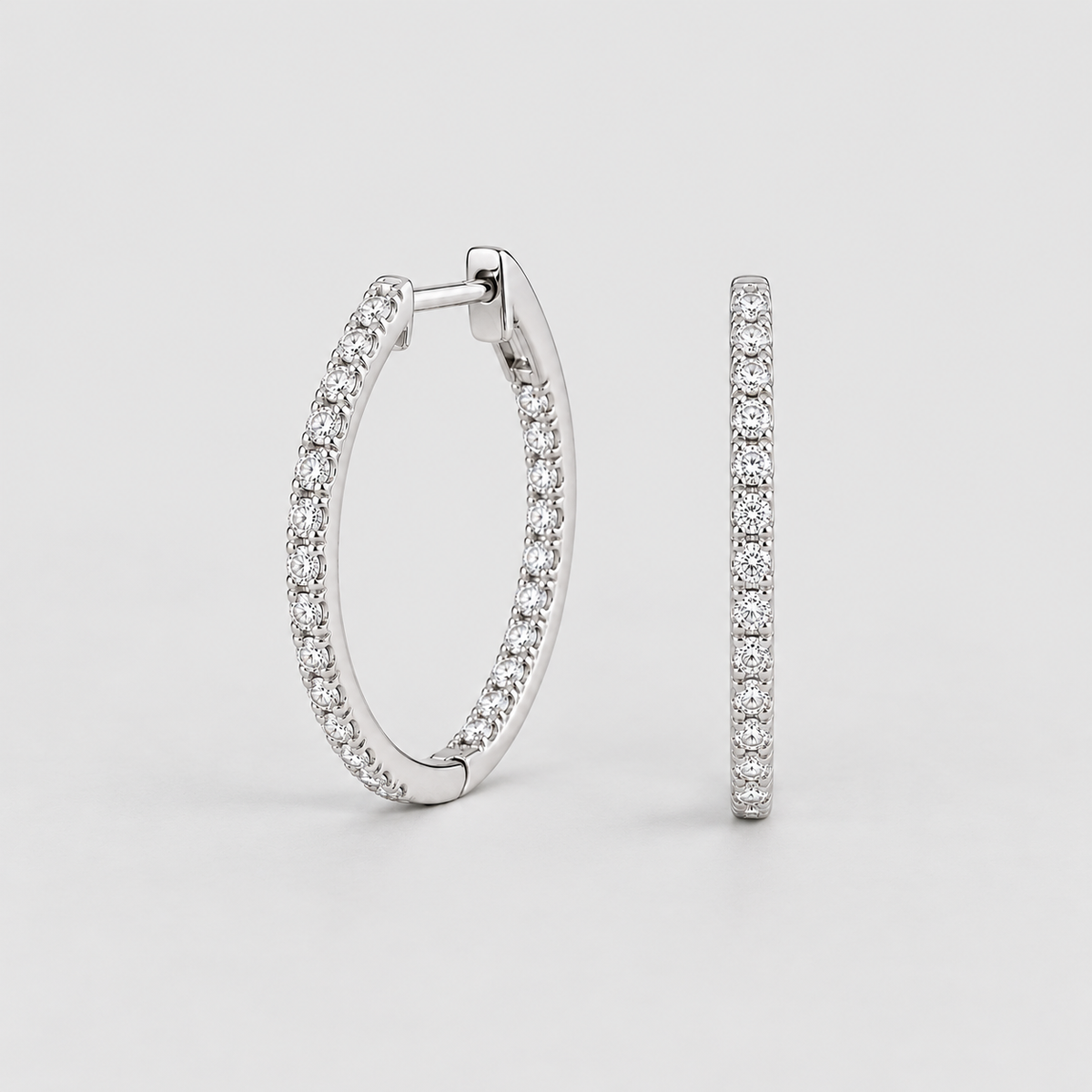 A pair of silver hoop earrings paved with small clear stones, shown from the front and side against a light gray background.