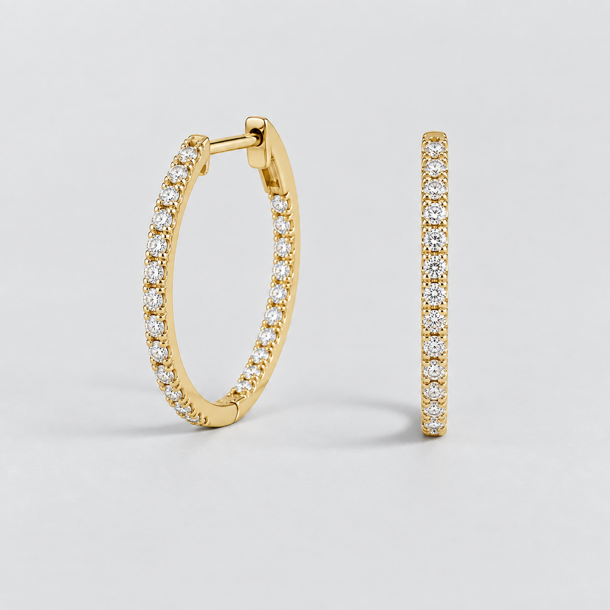 A pair of gold hoop earrings, one shown from a three-quarter angle and the other from a side profile view, both adorned with rows of clear pavé-set gemstones, on a light gray background.