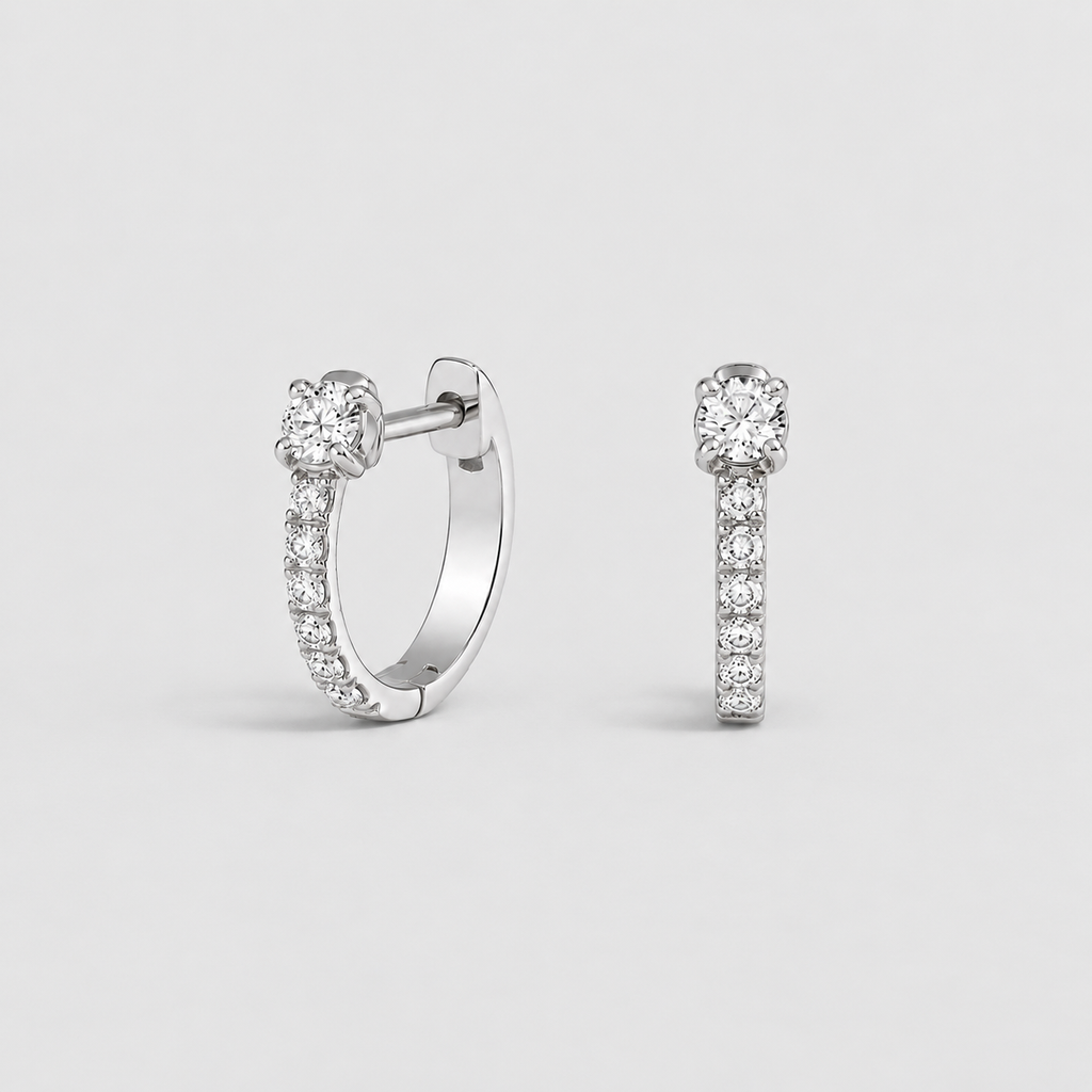 A pair of silver huggie hoop earrings, one shown from the front and the other from the side, both featuring a pavé-set diamond band and a central round-cut diamond, on a light gray background.