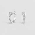 A pair of silver huggie hoop earrings, one shown from the front and the other from the side, both featuring a pavé-set diamond band and a central round-cut diamond, on a light gray background.