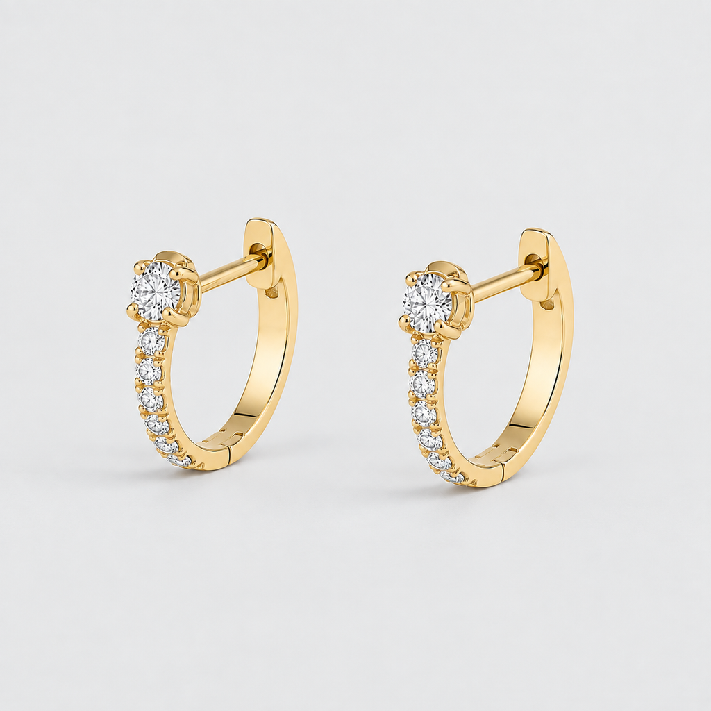 A pair of gold huggie hoop earrings with a single round-cut center stone and a row of small pavé-set clear stones along the front, shown on a light gray background.