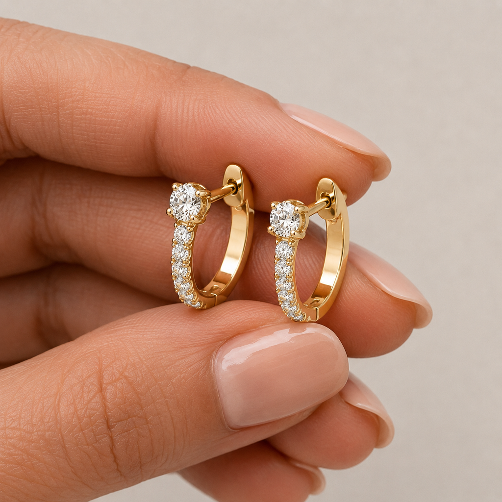A close-up of a hand holding a pair of small gold huggie hoop earrings with pavé-set clear stones.