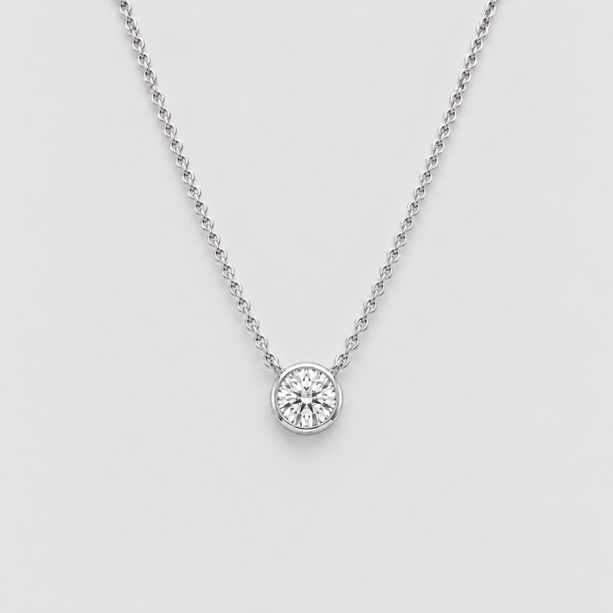 A delicate silver chain necklace with a single round bezel-set diamond pendant, shown against a clean light gray background.