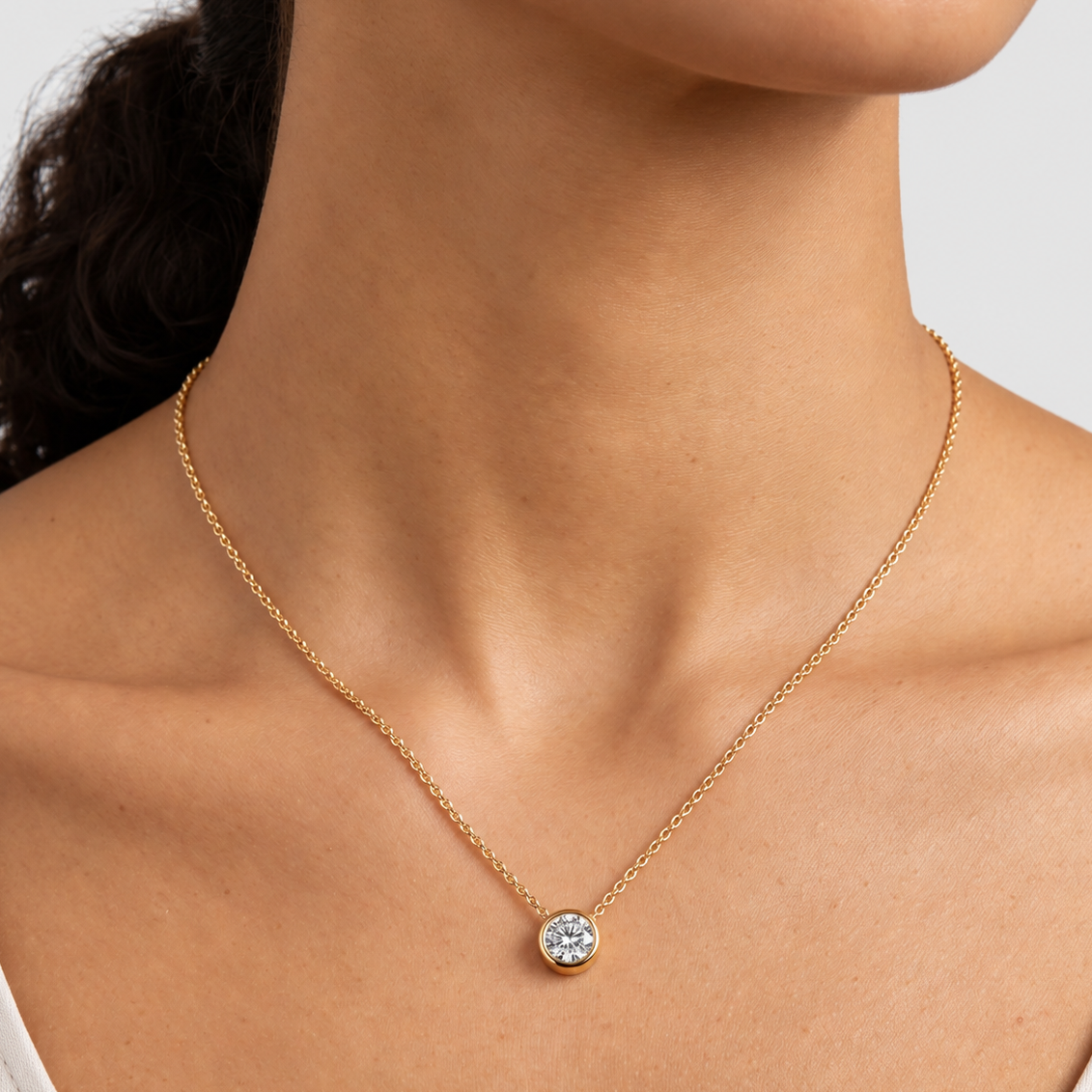 A close-up of a model's neck and upper chest wearing a delicate gold chain necklace with a small round bezel-set clear gemstone pendant.