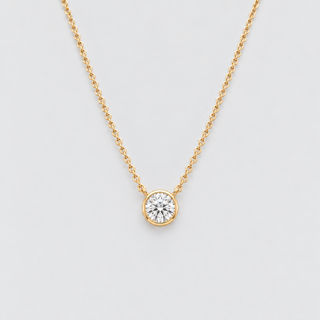 A delicate yellow gold chain necklace with a single round bezel-set diamond pendant, displayed against a clean light gray background.